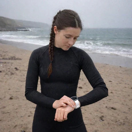 Female surfer wearing a white Garmin watch band over a wetsuit on a grey, drizzly UK beach