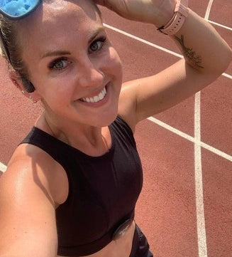 Woman smiling on an outdoor running track wearing a black sports top and a pink Apple Watch strap, taking a post-run selfie in the sun.