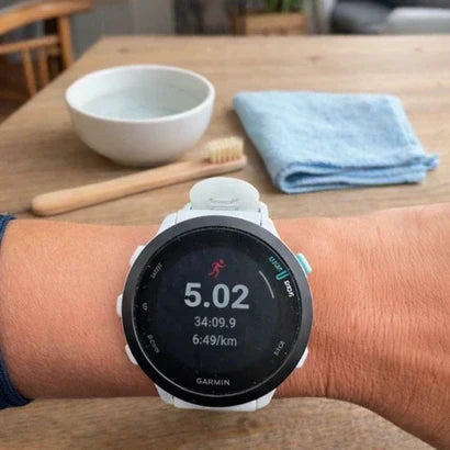 Garmin watch worn on the wrist with a white strap, placed above a wooden table with a bowl of water, soft cloth and cleaning brush.
