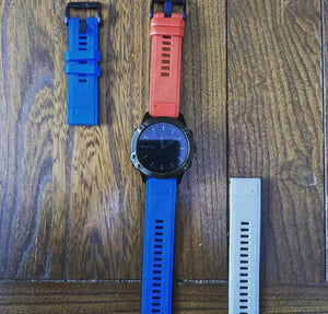 Garmin smartwatch on a wooden surface with interchangeable silicone straps in blue, red, and grey.